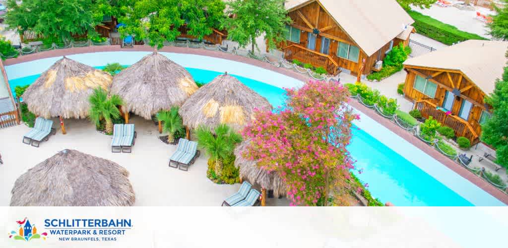 Aerial view of Schlitterbahn Waterpark and Resort featuring a serene pool flanked by loungers and thatched umbrellas, with lush pink flowers in bloom. Nearby cozy wood cabins offer a retreat aside the water paradise in New Braunfels, Texas.