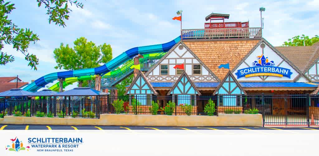 Image shows Schlitterbahn Waterpark and Resort in New Braunfels, Texas, on a sunny day. The facade has a traditional half-timbered design, with signage above the entrance. Behind, a large blue and green water slide curls around an elevated platform.