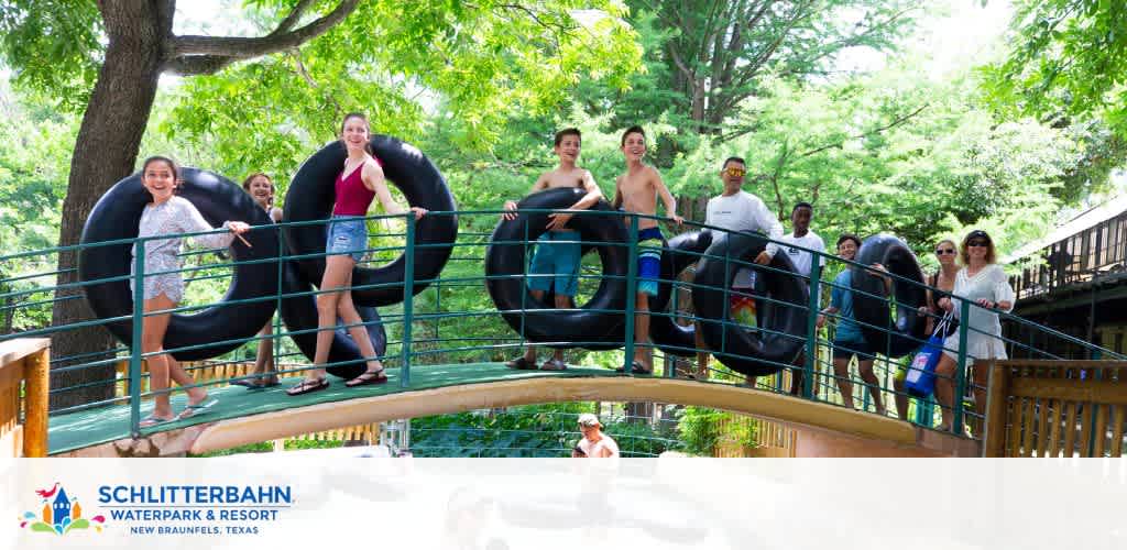 Guests at Schlitterbahn Waterpark and Resort in New Braunfels, Texas, are seen cheerful and ready for fun, holding black inner tubes and standing on a leafy, greenery-flanked walkway, with the park's logo and location displayed in the corner.