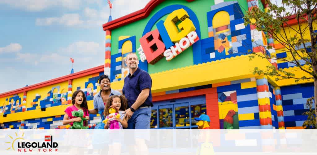 A family of four is smiling and walking hand-in-hand in front of a colorful building labeled LEGO SHOP at LEGOLAND New York. The facade resembles giant Lego bricks and the sky is bright and blue. They are holding stuffed toys and appear happy and excited.