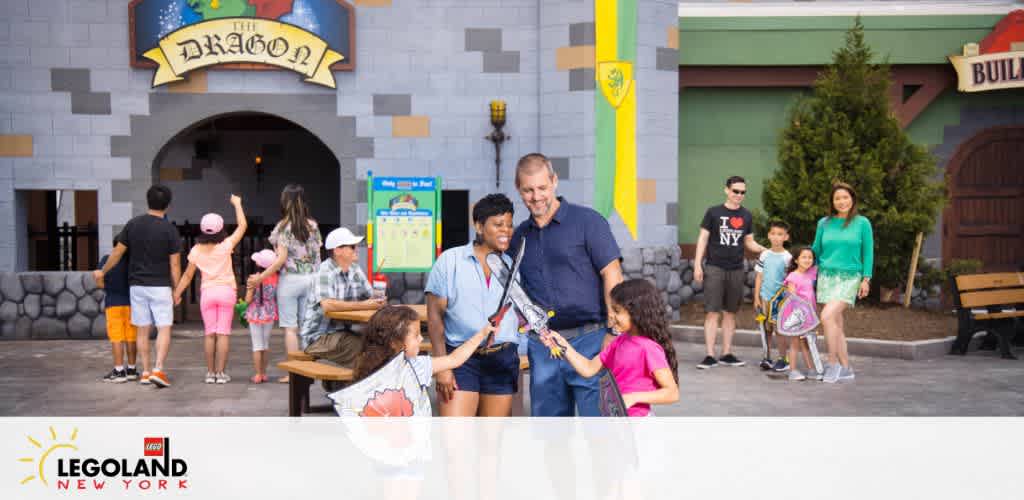 Visitors of various ages enjoy themselves outside 'The Dragon' ride at LEGOLAND New York. Smiling families are pictured standing and walking, with children holding LEGO toys. A stone-like archway with a colorful sign marks the entrance to the attraction, and the park's branding is visible in the corner.