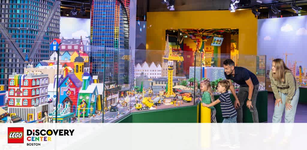 Image displays LEGO Discovery Center in Boston with vibrant LEGO cityscapes, featuring skyscrapers and colorful buildings. A family with two adults and two children are enjoying the elaborate displays, pointing and looking closely at the intricate details.