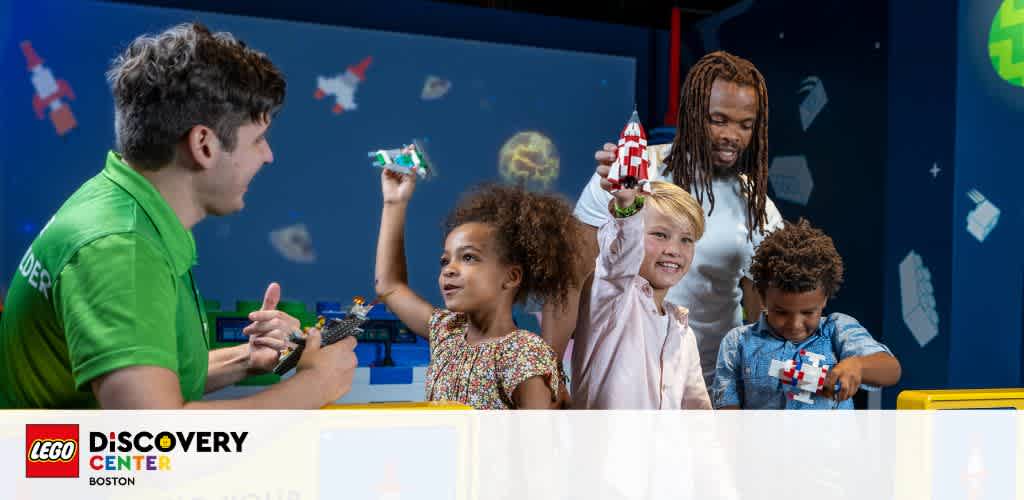 An instructor in a green shirt interacts with smiling children building Lego at the Lego Discovery Center Boston. A globe decoration is in the background, enhancing the playful learning atmosphere.