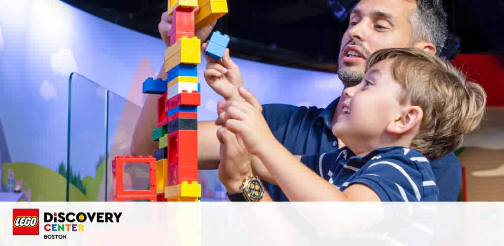 A man and a child are engaging in a playful activity, assembling a colorful structure with interlocking bricks. The setting is an indoor play area, as indicated by the signage in the background, promoting a family-friendly environment.