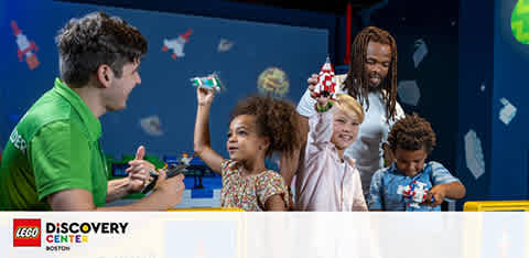 An engaging scene at the LEGO Discovery Center where an enthusiastic staff member interacts with three children and an adult. They are all focused on colorful LEGO bricks. The background features whimsical wall decorations, creating an atmosphere of creativity and fun.