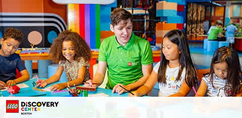 An instructor in a green shirt happily engages with three children at a colorful table within the LEGO Discovery Center. They are each focused on creative play and assembly tasks with LEGO pieces, in a bright, vibrant room full of playful elements and decor.