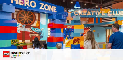 Image displays an indoor LEGO play area at the LEGO Discovery Center Boston. Visitors, including a man and two children, are observing colorful LEGO structures with signage that reads 'HERO ZONE' and 'CREATIVE CLUB.' The area is vibrant with a playful design.