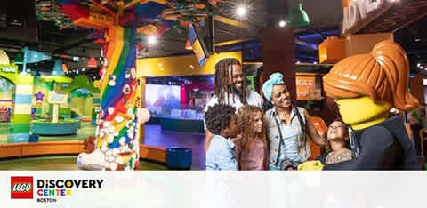 A lively scene at LEGO Discovery Center with a group of people, including children, interacting with a life-sized LEGO figure. Brightly colored play areas and structures fill the background, suggesting a fun and engaging atmosphere.