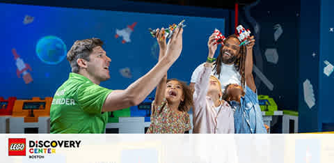 The image shows a joyful moment at the LEGO Discovery Center with two kids and two adults. The man on the left, wearing a green LEGO builder shirt, excitedly lifts a LEGO creation with a child next to him. On the right, a man lifts a happy child in the air, both holding up LEGO figures. The background includes a vibrant blue wall with LEGO-themed decorations. The LEGO Discovery Center logo is at the bottom left.