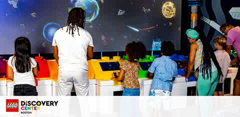 Visitors of varied ages are engaging with interactive exhibits at the LEGO Discovery Center. A large screen displays vibrant images of space, captivating the audience. The logo in the corner signifies the playful and educational environment the center provides.