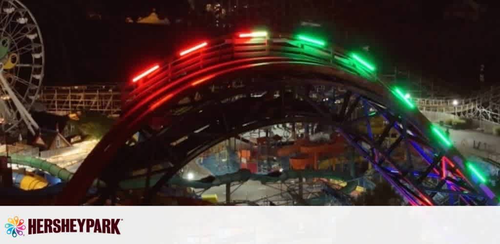 This image captures an illuminated roller coaster track at Hersheypark during nighttime. The track glows with vibrant red, green, and blue lights, curving against a backdrop of other park rides and attractions. Below, the festive lights from the park are visible, and the structure appears to be active and lively. The Hersheypark logo is displayed at the bottom left.