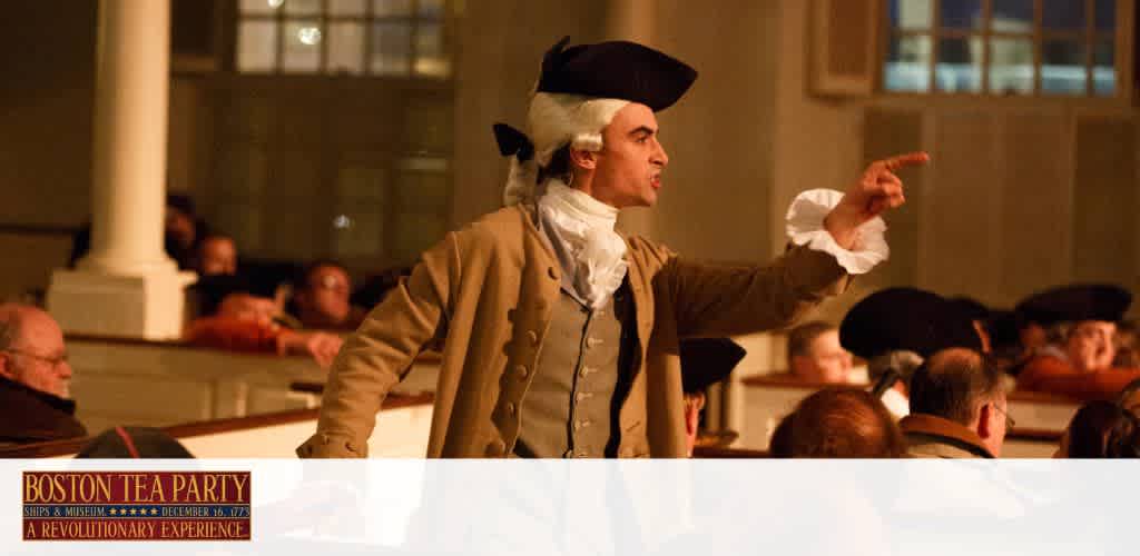 A historical reenactor dressed in 18th-century attire, including a tricorn hat, is gesturing passionately during a speech at the Boston Tea Party Ships & Museum. The setting is dimly lit to evoke the era, with a captivated audience in colonial dress seated in the background. The lower part of the image features the text 'Boston Tea Party - A Revolutionary Experience'.