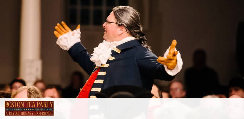 Image shows a person dressed in historical attire with a navy coat, white ruffles at the wrist, and a tricorner hat. They are gesturing passionately with both hands raised. A banner at the bottom reads 'Boston Tea Party Ships & Museum - A Revolutionary Experience'. The setting appears to be an indoor event with dim lighting and an audience in the background.