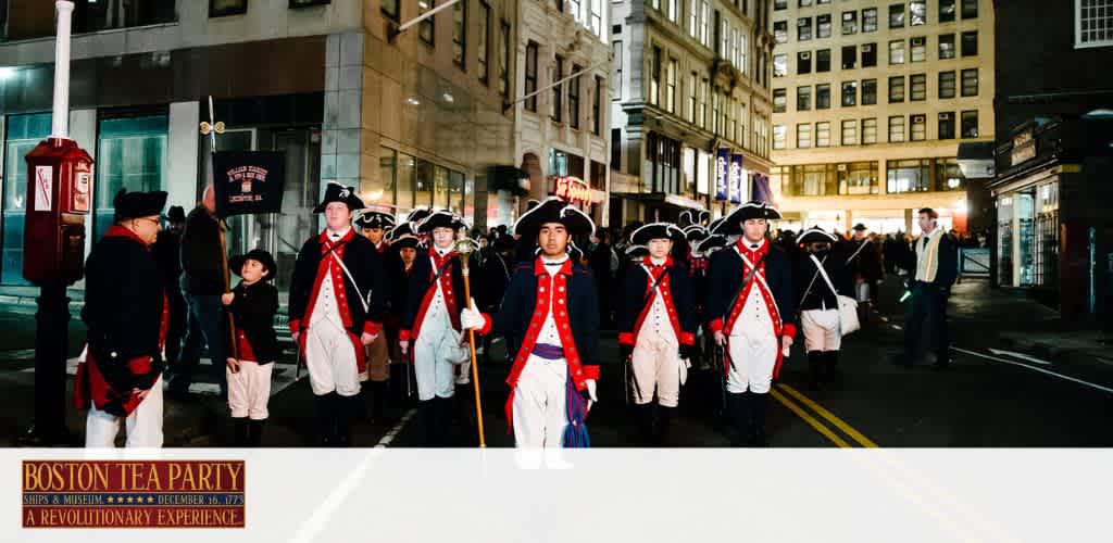 The image features a group of individuals dressed in historical costumes resembling those of the American Revolutionary period, marching along a city street at night. They are wearing red and blue coats with white breeches and black tricorn hats, carrying muskets. A sign on the left advertises the Boston Tea Party Ships & Museum event, hinting at a reenactment or historical celebration.