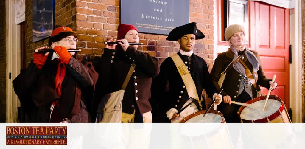 Image of a historical reenactment at the Boston Tea Party Ships and Museum. Four individuals in colonial-era clothing stand against a brick wall background. From left to right, two are playing flutes, and the other two are playing drums. An informational sign mentioning the historic event is partially visible in the background. The ambiance evokes the spirit of a bygone era.
