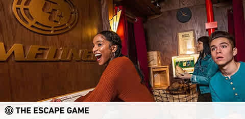 Image Description: This is a vibrant and dynamic photo from "The Escape Game" venue. In the forefront, there are two participants engaged in the game. On the left, a person with a joyful expression is looking directly at a clue or object, showing a look of excited discovery. This individual is wearing a ribbed, rust-colored sweater. To the right, another player with an astonished expression is looking upward, possibly at a clue or element out of frame, wearing a teal button-down shirt. Both are surrounded by a well-themed room with wood panel walls, antiques, and various props that create an immersive experience, suggesting a puzzle-solving atmosphere. Above them, a large wooden sign with the venue's name is prominently displayed. At FunEx.com, we're committed to offering you the best experience with exclusive savings – guaranteeing you the lowest prices on tickets for your next thrilling escape!