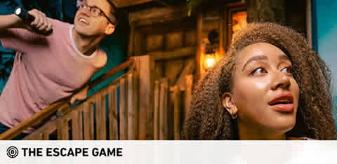 This image showcases two individuals engaged in an interactive experience at The Escape Game. On the left side of the picture, a man with glasses, wearing a casual t-shirt, extends his arm out as if he’s reaching for something or pointing while looking excited and focused. He is standing on a set of wooden stairs which add to the rustic and adventurous atmosphere. On the right, a woman is closer to the foreground, looking upwards with an expression of thoughtful curiosity. She has curly hair and seems to be pondering a clue or deciding her next move within the game scenario. The lighting in the image is warm, suggesting an indoor setting with a hint of mystery.

At GreatWorkPerks.com, we're committed to finding you the lowest prices on tickets so you can enjoy immersive experiences like The Escape Game without breaking the bank.