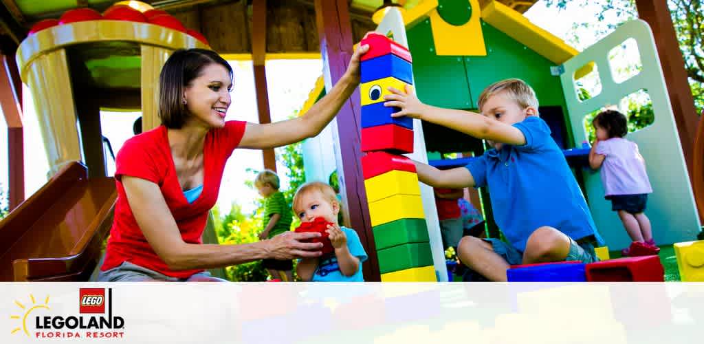 A joyful scene at LEGOLAND Florida Resort with a woman smiling as she interacts with a toddler. A boy in a blue shirt plays with oversized colorful Lego blocks, focused on building a tall structure, while other children are seen engaged in play in the background of a vibrant play area.