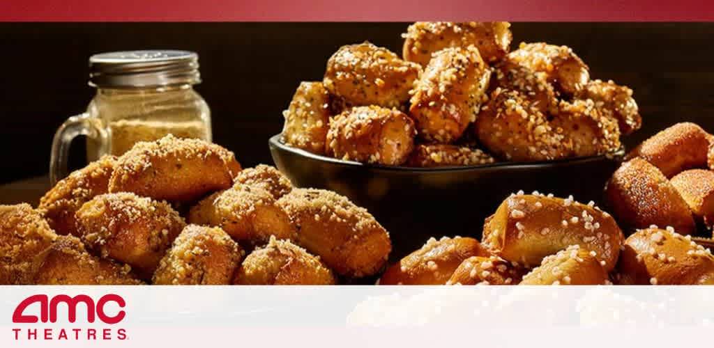 Image Description: This is a promotional image featuring an appetizing display of food associated with AMC Theatres, which is indicated by the logo at the bottom left of the picture. In the foreground, there's a large platter filled with golden brown pretzel bites generously coated with sesame seeds. The bites have a glistening appearance, suggesting they are freshly baked and possibly buttered. Behind the platter, a glass mason jar is visible, which might typically be used for serving or storing beverages or condiments. The background is dark, focusing the attention on the pretzel bites and creating a warm, inviting atmosphere typical of a movie theater setting.

End with promotional sentence: "Experience the perfect snack to complement your movie-going experience, and don't forget to check GreatWorkPerks.com for the lowest prices on tickets and great savings on your next AMC Theatres visit!"