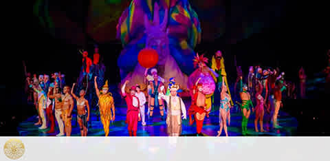 Vibrant theatrical performance featuring a diverse cast of performers in colorful costumes, posing on stage. There's a festive atmosphere with a whimsical backdrop styled as an oversized, playful creature, adding to the magical feel of the scene.