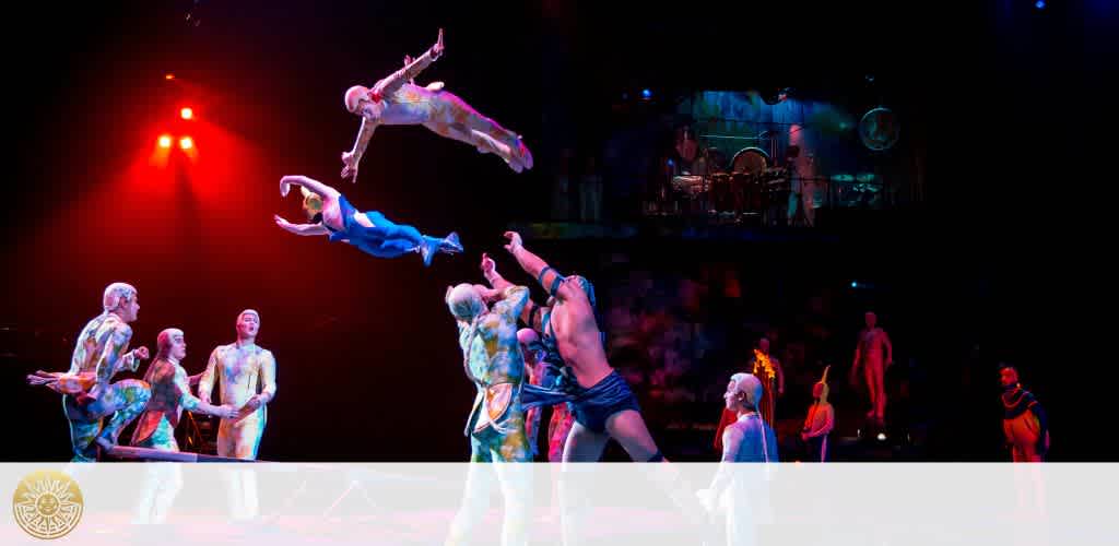 Vividly colored image from a circus performance showing acrobats mid-air after being launched by their partners. Two acrobats are flying above the stage, one in a blue costume, the other in white. Below them, performers in vibrant, patterned body suits watch intently. A drummer on an elevated platform in the background adds to the dynamic scene.