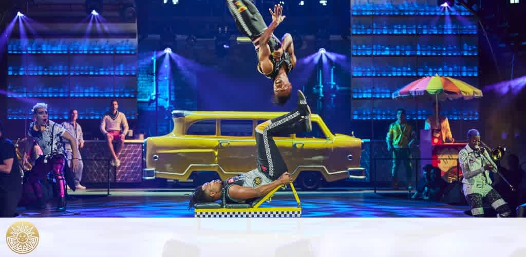 On stage, a performer executes a backflip over another who's lying down, while musicians, including a saxophonist, play by a yellow taxi and beach umbrella. The setting has vibrant lighting with a blue backdrop. The Cirque du Soleil logo is visible.