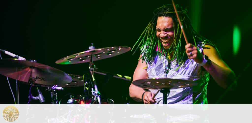 A person with dreadlocks is joyfully playing the drums on stage, surrounded by green lighting. The focus is on the drummer in motion, with drumsticks in the air and a delighted expression on their face.