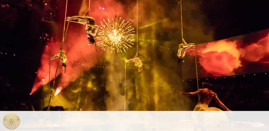 An energetic live performance with aerial acrobats suspended on ropes against a backdrop of vibrant fireworks and dramatic red-hued smoke. One performer is actively engaging with the audience on stage right.