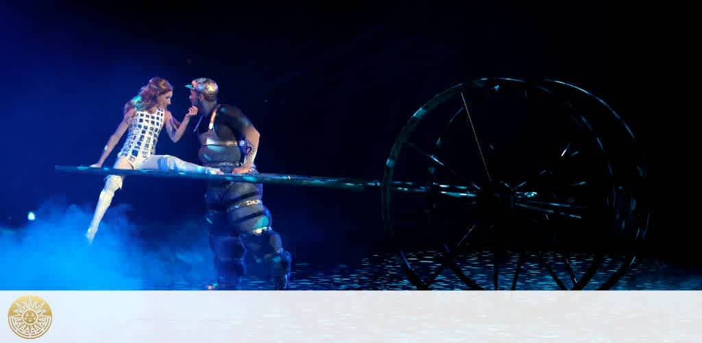 A stage performance is captured with two individuals mid-act. The person on the left balances elegantly on a beam, dressed in a white, patterned outfit. To the right, a performer in a dark, armored costume supports the beam. A large, dark wheel structure stands in the background, all under blue lighting. A logo with a leafy emblem is in the corner.