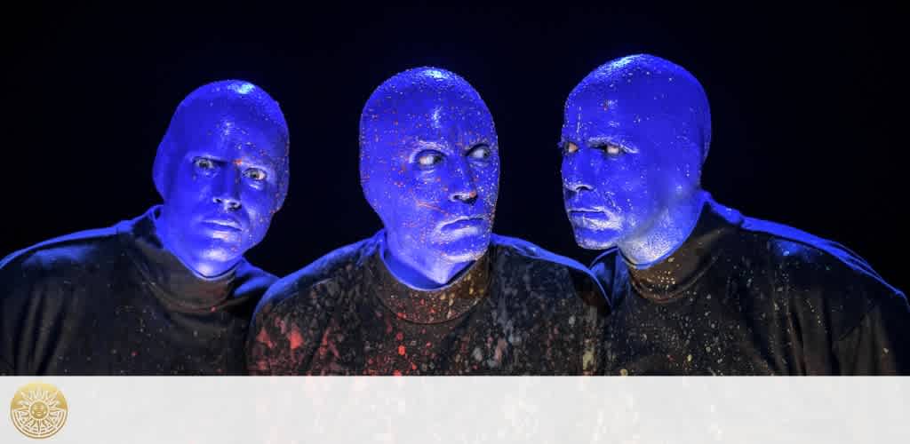 Image description: Three artists with blue-painted skin and bald heads are closely grouped together against a dark background. They wear expressionless faces and black clothing splattered with bright specks, appearing both intriguing and mysterious.