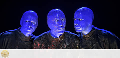 Image features three individuals with blue-painted faces and black clothing, standing against a black background. The lighting highlights their expressions of surprise and curiosity. They are reminiscent of a performance group known for visual spectacles. The image conveys a dramatic and artistic mood.