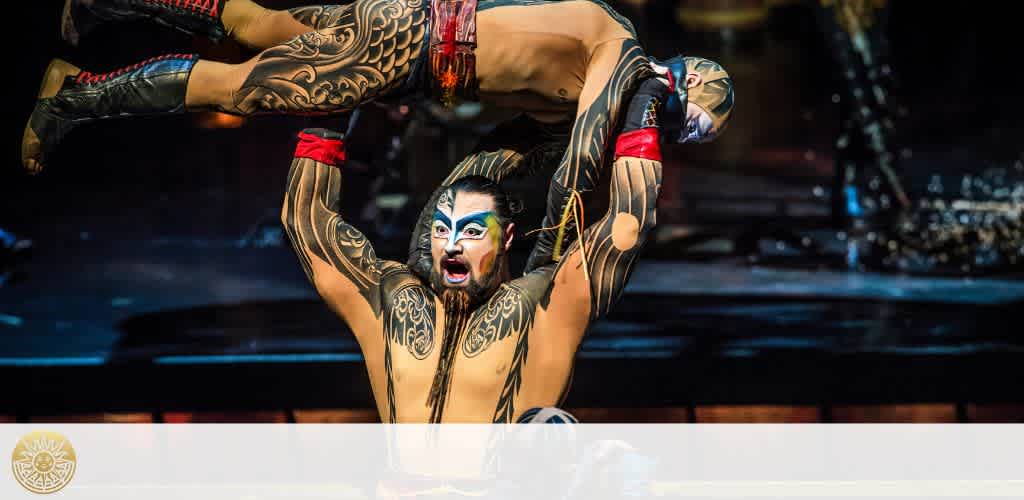 A performer with dramatic makeup and tribal-patterned body paint is captured mid-flip on stage, supported by another person's outstretched arms. They wear an elaborate costume with red accents. The backdrop is dimly lit, focusing attention on the action.