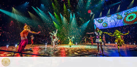 Image of a vibrant stage performance with performers in colorful costumes under bright lights. Confetti flies through the air as one artist balances on a wire, and others engage in dynamic dance and acrobatics. The background features a large screen with eye-catching patterns. A festive and lively atmosphere is conveyed through the visual spectacle.