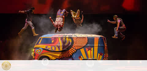 A vibrant performance scene with four acrobats in mid-air above a colorfully painted van with patterns resembling animal motifs. The background is a warm red, with mist effect near the van, suggesting dynamic movement.