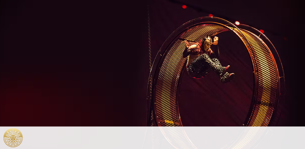 This image depicts a captivating circus performance featuring a performer dressed in an elaborate costume with reflective elements, suspended mid-air inside a large, luminous mechanical wheel. The wheel is adorned with a series of red lights along its circumference, which stand out against the dimly lit, dark background of the stage. The performer is caught in a dynamic pose with limbs outstretched, conveying a sense of motion and gravity-defying skill. To the bottom right, there is a logo consisting of a sun with decorative rays and a prominent crest-like emblem in its center.

At GreatWorkPerks.com, we pride ourselves on offering the thrill of live entertainment at the lowest prices. Find the best deals and savings on tickets to astonishing performances like this when you book with us.