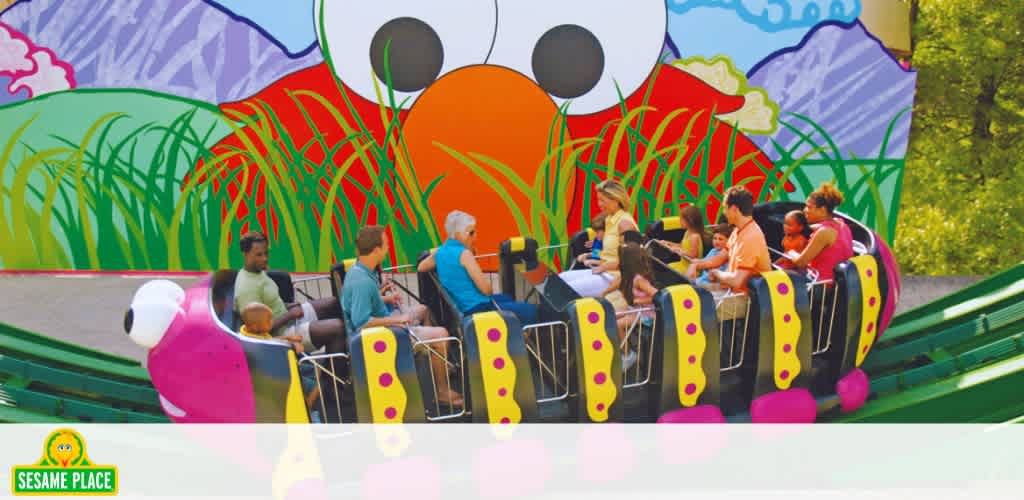 Image shows visitors enjoying a ride at Sesame Place. The ride car is green and yellow, resembling a caterpillar. Colorful background illustrations depict scenery and the iconic Sesame Street character Elmo's face. The Sesame Place logo is visible in the corner. Visitors of various ages are seated, appearing engaged and happy.