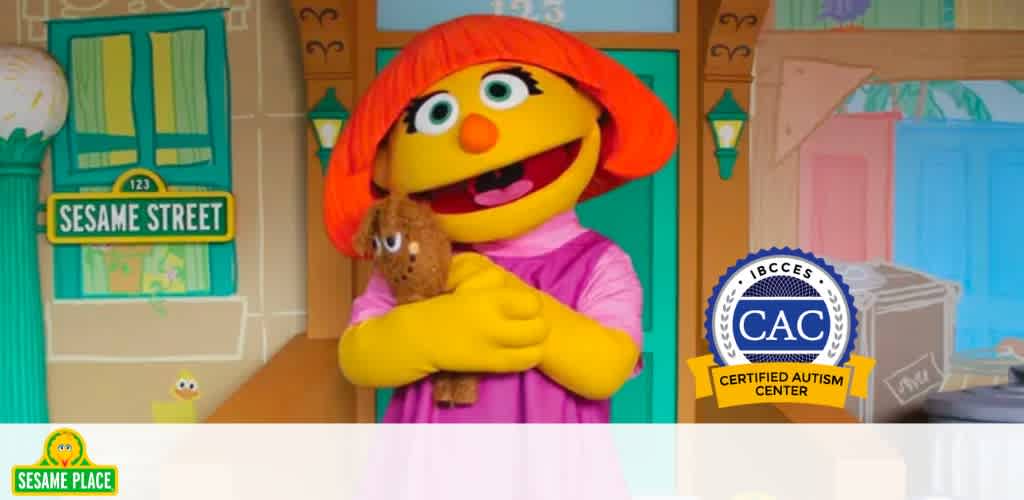 An image featuring a smiling orange Muppet with yellow hair and a pink dress, hugging a brown teddy bear. In the background, colorful cartoon-style house facades display the text 'Sesame Street' and house number '123'. In the corner, logos for 'Sesame Place' and a 'Certified Autism Center' seal are visible.
