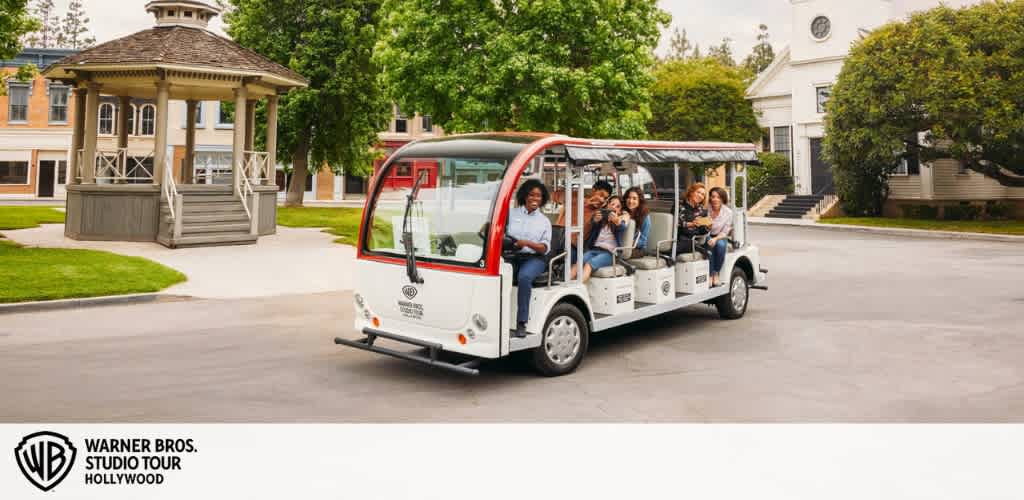 Image shows a group of joyful visitors on an open-sided shuttle during the Warner Bros. Studio Tour Hollywood. They are passing by a gazebo and classical buildings on a sunny day, waving and enjoying the tour. The Warner Bros. logo is visible in the corner.