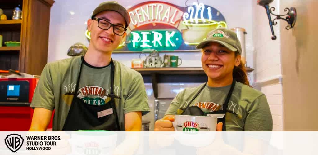 Image shows two cheerful employees wearing green Central Perk t-shirts and aprons, serving coffee inside the recreated set of a famous TV show coffeehouse, with its iconic neon logo in the background. The Warner Bros. Studio Tour Hollywood logo is visible.