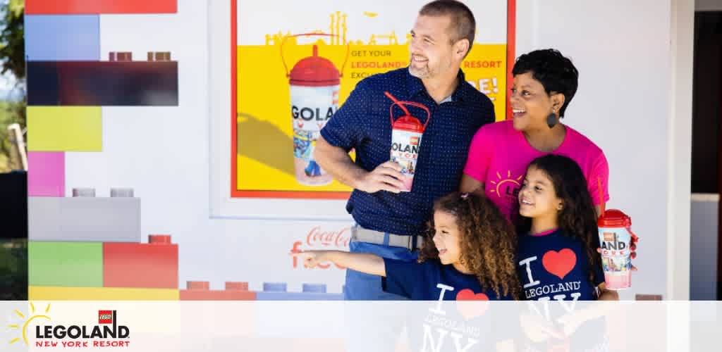 A happy family of four stands with Legoland New York Resort drinks. Two adults and two children smile, wearing shirts that express their love for Legoland. A colorful Legoland promotional background enhances the joyous atmosphere. They all seem to be having a great time.