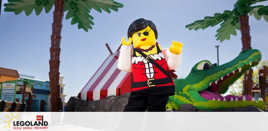Image shows a vibrant scene at LEGOLAND New York Resort with a life-sized, brick-built figure resembling a pirate, complete with a black hat, eye patch, and red coat, holding a treasure map. In the background, a large, playful LEGO alligator peers out from the water, surrounded by tropical LEGO palm trees and brightly colored buildings.