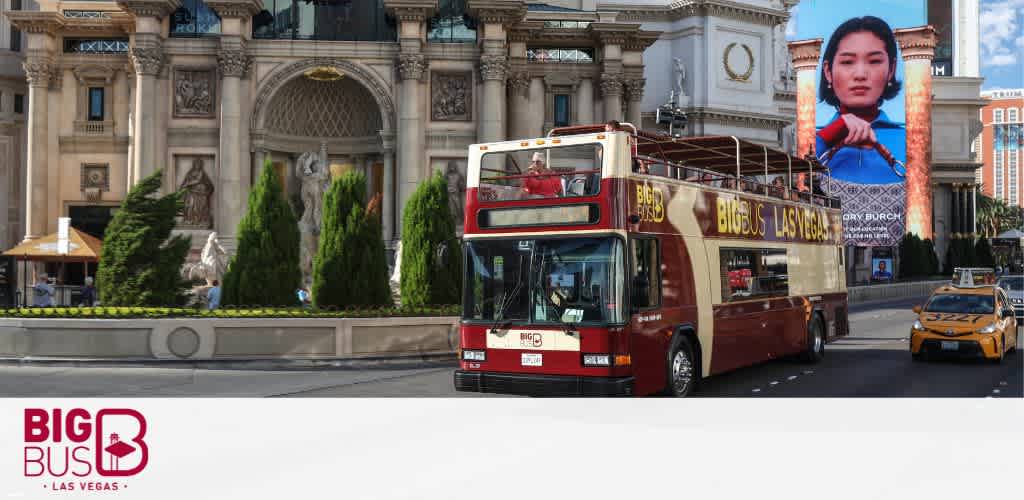 Image shows a red open-top tour bus labeled 'Big Bus Las Vegas' driving on a street in a sunny setting. A yellow taxi and grand architecture, including ornate statues and digital billboards, are visible in the background, indicative of a vibrant city scene.