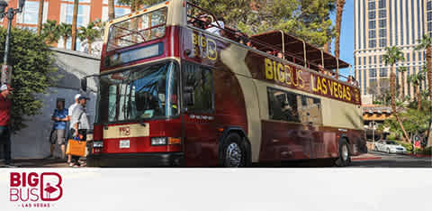 A red, open-top double-decker Big Bus in Las Vegas is parked with people boarding. Palm trees and a clear sky are visible in the background with urban buildings. The bus features prominent branding and the destination on the front display.