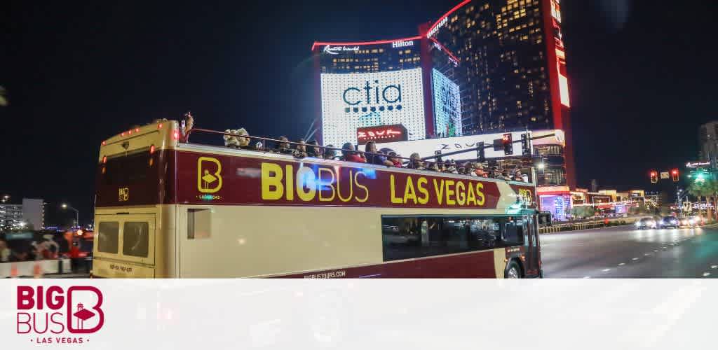 A Big Bus Las Vegas tour bus with open-top seating is shown at night. Passengers are visible on the upper deck, enjoying the brightly lit cityscape, which includes illuminated buildings and billboards. The bus and the vibrant backdrop emphasize the lively experience of touring Las Vegas after dusk.