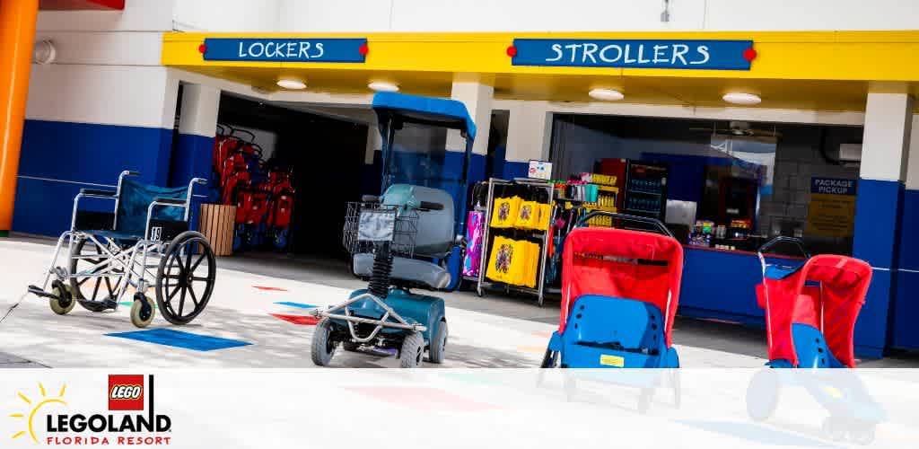 Image of a facility at LEGOLAND Florida Resort offering lockers and strollers. Visible are wheelchairs and strollers available for rent, and lockers for storage, showcasing the accessibility options for visitors with different needs. The area has distinct blue and red color themes with clear signage.