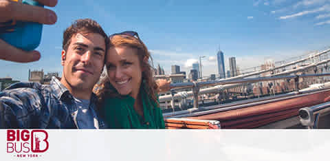 Image shows a smiling man and woman taking a selfie on a Big Bus New York tour. They're seated, and the city skyline, including a tall skyscraper, can be seen in the background under a clear blue sky. The Big Bus logo is visible in the corner.