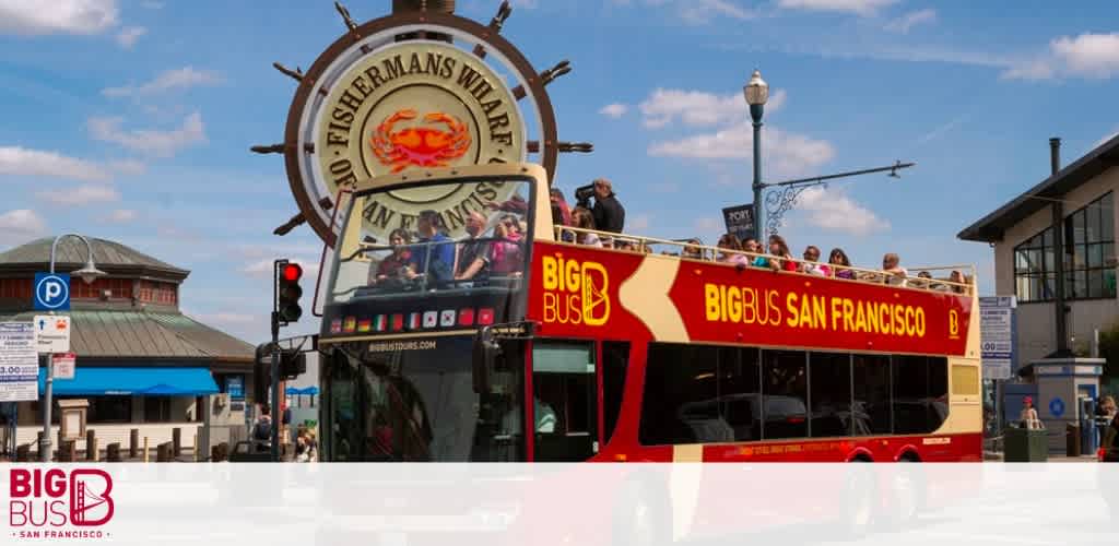 Image shows a red open-top tour bus branded with 'BIG BUS SAN FRANCISCO' on its side, carrying passengers. In the background, a large sign reads 'Fisherman's Wharf' under a clear blue sky. The setting suggests a sunny day, perfect for sightseeing in San Francisco.