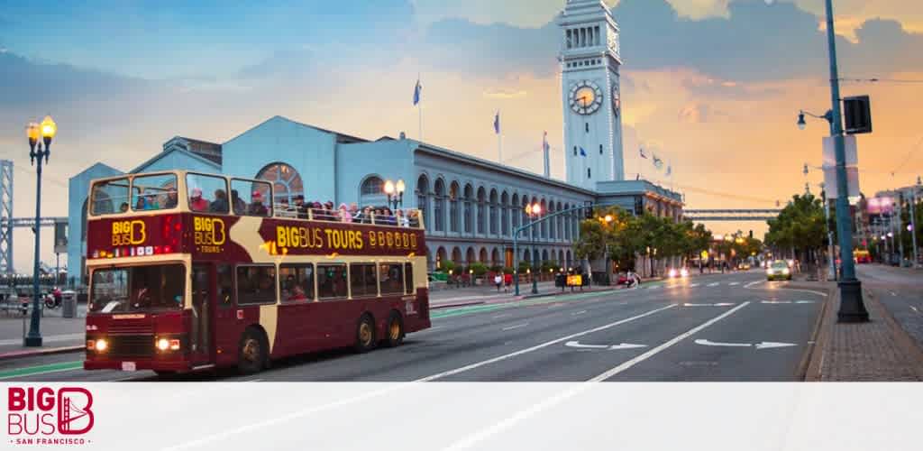 A red double-decker tour bus with the text 'Big Bus Tours' on its side moves along a city street at dusk with the illuminated Ferry Building clock tower in the background. The sky is tinged with soft blue hues, signaling evening, and street lamps cast a warm glow along the bustling road.