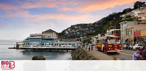 Sunset over a waterfront with buildings, a tour bus on the street, and hills in the background.