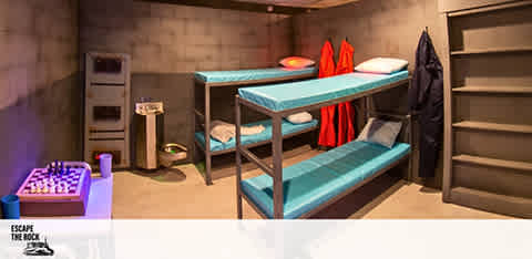 Image Description: This is an interior view of a themed escape room designed to resemble a bunker. The floors and walls are solid gray, simulating a reinforced concrete look typical of bunkers. On the left side, there's a purple chess board set on the floor, ready to be played. The center of the room features a sturdy blue bunk bed with three levels, each outfitted with a neatly arranged red towel and a gray pillow. To the right, a row of metal lockers stands beside the bed with the door of one locker ajar, giving a hint of an interactive element to the space. Against the back wall, two round, red-backed chairs sit in expectation of participants. The lighting is soft, creating a secretive atmosphere that underscores the room's escape challenge theme. On the bottom left corner, text reads "ESCAPE THE ROOM," suggesting the purpose of this engaging environment.

While exploring exciting experiences to add to your adventure list, remember that at GreatWorkPerks.com, you can find the lowest prices and amazing savings on tickets to a vast array of entertainment options.